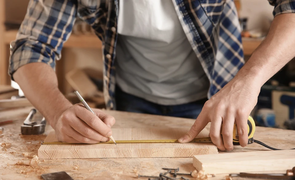 Homme souriant dans atelier de menuiserie tenant une planche de bois, entouré d'outils de travail du bois